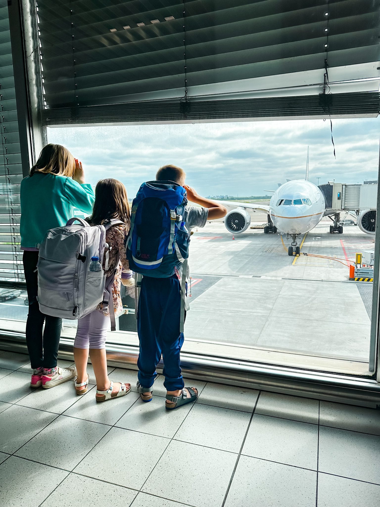 three kids looking out of a window of an airport at an airplane. They're wearing backpacks great for traveling.